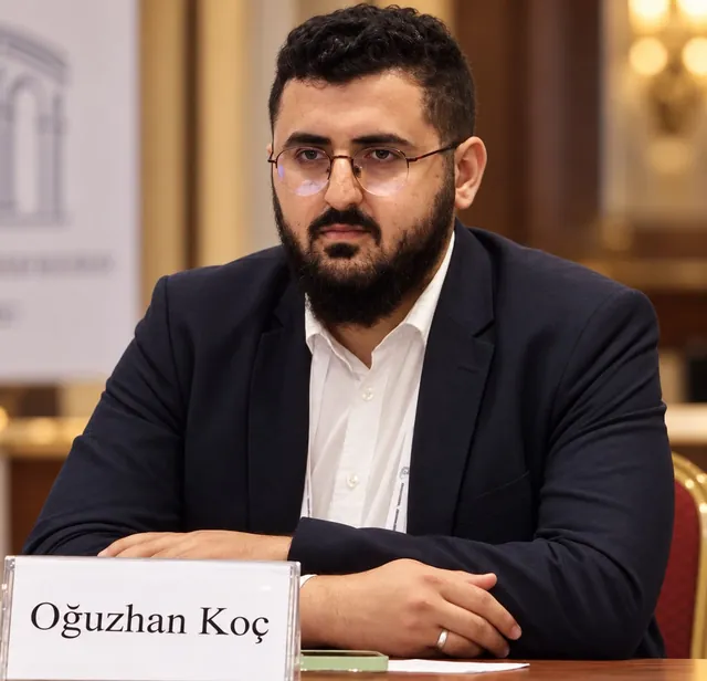 Oğuzhan Koç’un portresi: gözlüklü, sakallı, lacivert ceketli; bir toplantı masasında oturuyor